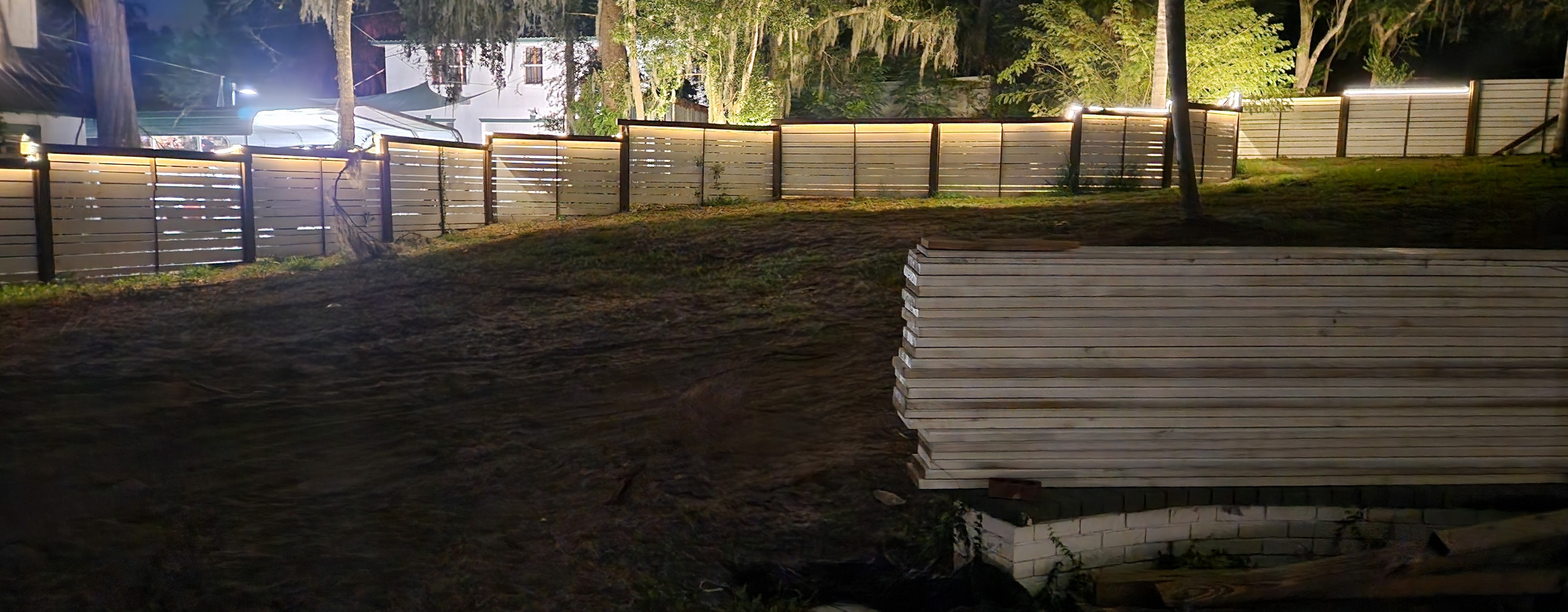 Illuminated horizontal fence installation at night with lumber staged and ready for the next phase of construction.